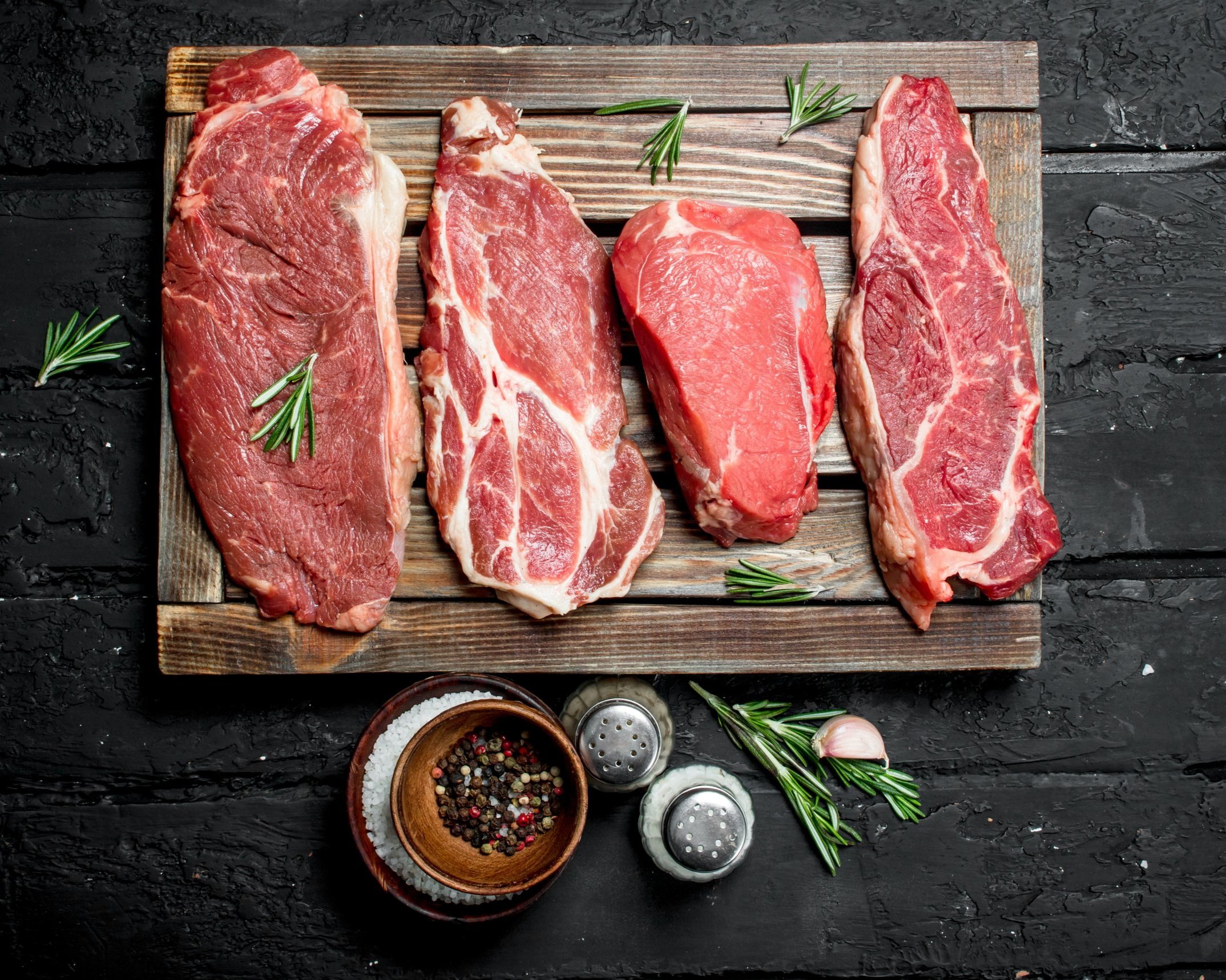 Fresh cuts of beef displayed on a wooden board, featuring various steak types alongside sprigs of rosemary and seasonings in bowls, set against a textured black background. Ideal for meat lovers and culinary enthusiasts.