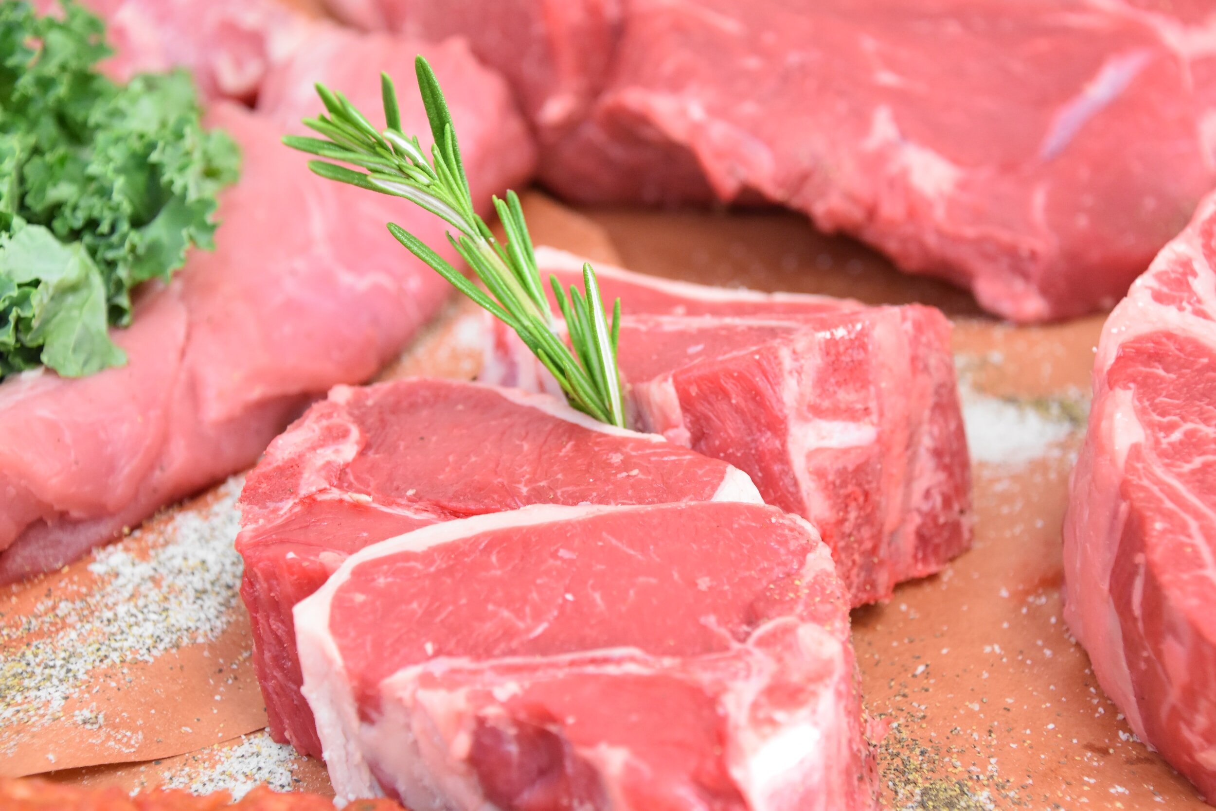 Fresh cuts of red meat arranged on butcher paper, featuring a sprig of rosemary and a side of leafy greens, ideal for cooking or grilling.